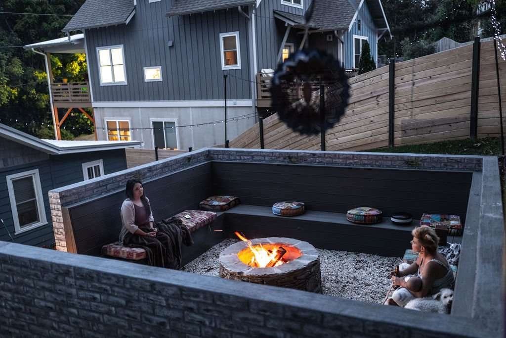 Laughter Family Hardscapes Near Me Asheville NC (6) Cozy outdoor fire pit area with two women seated on benches, surrounded by landscaped walls and decorative cushions, showcasing a relaxing hardscaping design by Laughter Family Hardscapes in Asheville, NC.