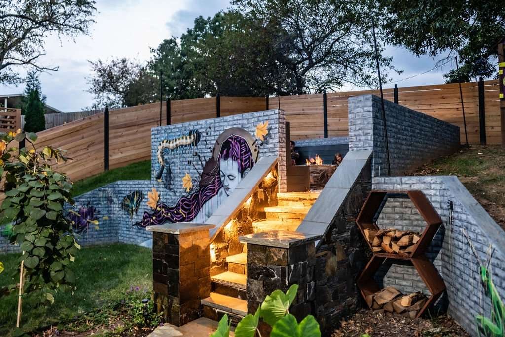 Laughter Family Hardscapes Near Me Asheville NC (5) Outdoor hardscaping featuring a stone staircase with illuminated steps, vibrant mural, and wooden fencing, enhancing a backyard space for family gatherings and relaxation.