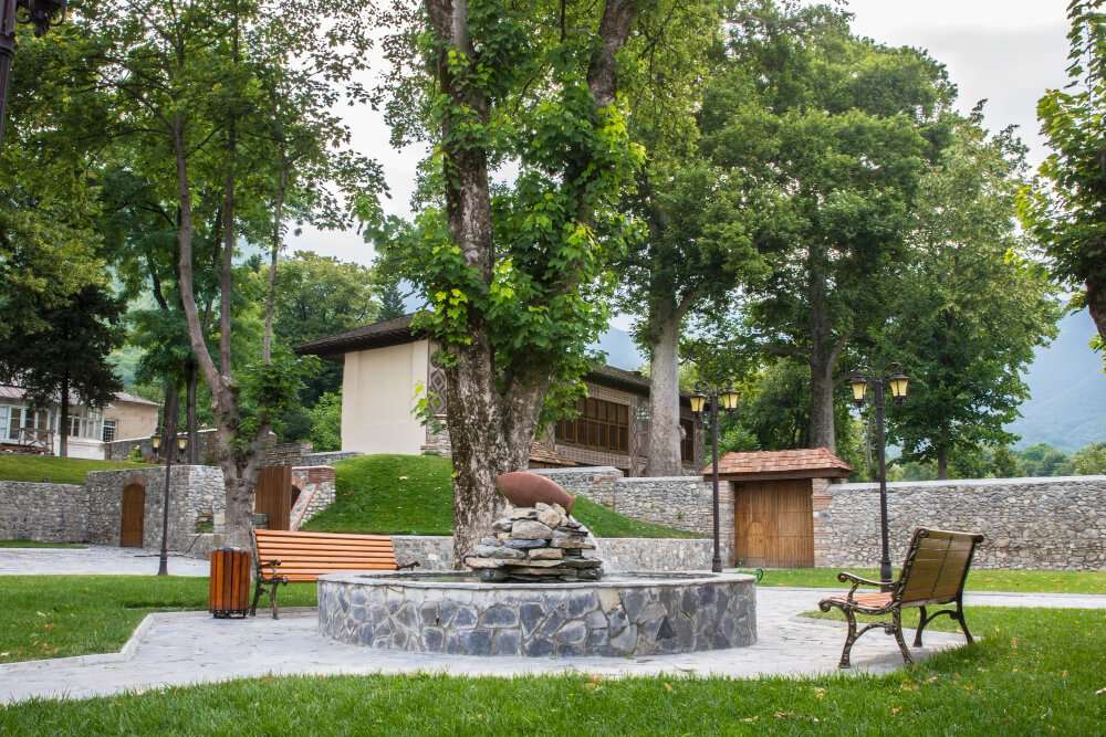 Stone fountain surrounded by benches and trees in a landscaped garden, enhancing outdoor space aesthetics for residential hardscaping.